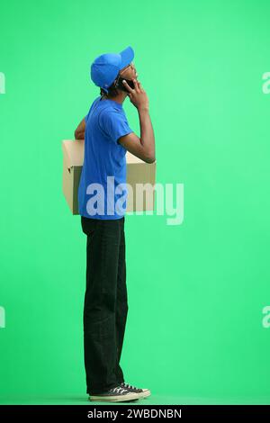 Ein männlicher Zustellmann, auf grünem Hintergrund, in voller Höhe, mit einer Box und einem Telefon Stockfoto