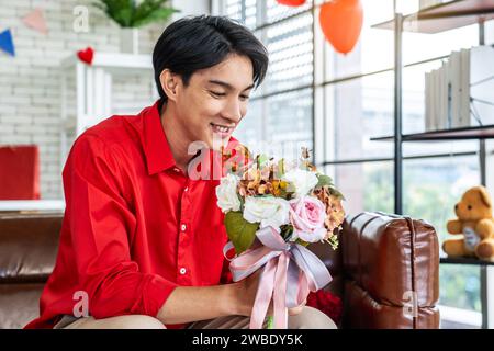 Junger asiatischer Mann mit einem Blumenstrauß in den Händen, der am Valentinstag auf dem Sofa in seinem Zimmer sitzt und eine Überraschung für seine Freundin vorbereitet. Stockfoto