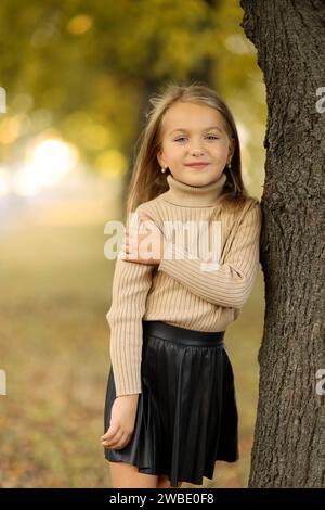 Kindertag. Fröhliches kleines Mädchen 5-6 Jahre alt posiert und lächelt draußen vor der Kamera und steht am Baum im Herbstpark. Stilvoller Zartpressdruck Stockfoto