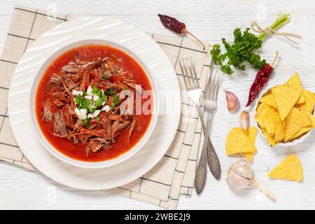 Birria de res, mexikanischer Rindereintopf in heißer Paprikasauce mit roher Zwiebeln und gehacktem Koriander in weißer Schüssel auf weißem Holztisch mit Taco-Chips und mir Stockfoto