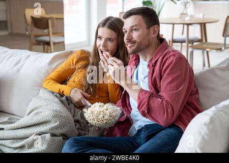 Interessierte Männer und Frauen essen Popcorn und schauen sich die Fernsehsendung an, die auf dem Sofa sitzt und Popcorn isst. Stockfoto