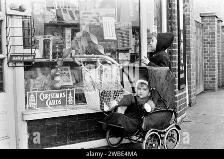 Ein Baby in einem Kinderwagen und ein Junge, der in das Fenster vor Souch's Zeitungskiosk blickt, Tabak & Post Office, Lordship Lane, Tottenham, 8. Januar 1969 Stockfoto