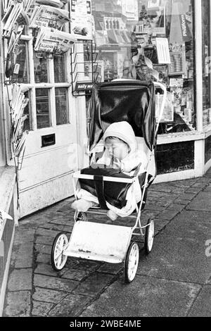 Ein Kleinkind, das in einem Kinderwagen vor Souch's Zeitungskiosk, Stationer & Post Office, Lordship Lane, Tottenham, London, UK, gelassen wurde - Mittwoch, 8. Januar 1969 Stockfoto