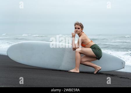 Das Sportmodemodell hält das Surfbrett und sitzt davor und blickt mit Nachsicht in die Distanz vor dem Hintergrund der Meereswellen. Wunderschön W Stockfoto