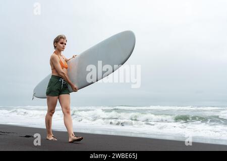 Surferin spaziert am schwarzen Sandstrand mit weißem Surfbrett vor dem Hintergrund der Meereswellen während der Sommerferien. Sportliche Frau im Bikinioberteil Stockfoto