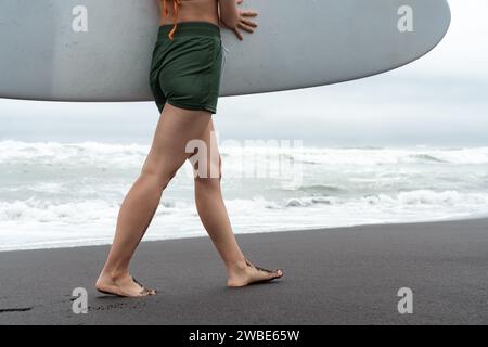 Surferfrau mit Surfbrett, die am Strand entlang läuft, mit Meereswellen im Hintergrund. Seitenansicht der Sinnlichkeit weiblicher Beine und Gesäß Stockfoto
