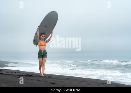 Surferin in in Bikinioberteil, Shorts und barfuß am schwarzen Sandstrand und mit weißem Surfbrett auf dem Kopf vor dem Hintergrund der Meereswellen Stockfoto
