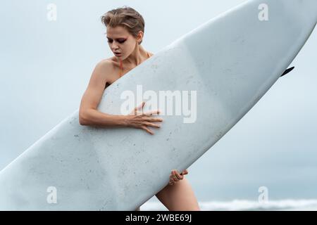 Nahaufnahme eines weiblichen Sportmodemodells, das ein weißes Surfbrett hält und dahinter steht. Surferin in in entspannter Pose, die nach unten blickt Stockfoto