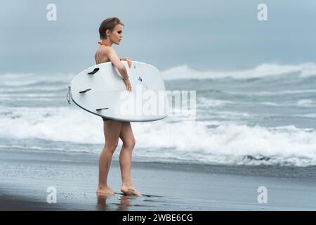 Niedliche Surferin Frau, die auf schwarzem Sandstrand steht und während der Sommerferien weißes Surfbrett vor dem Hintergrund der Meereswellen trägt. Sinnlichkeit Frau Stockfoto