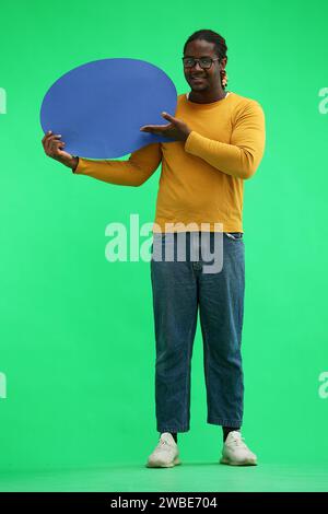 Ein Mann in einem gelben Pullover auf grünem Hintergrund hält ein blaues Kommentarzeichen Stockfoto