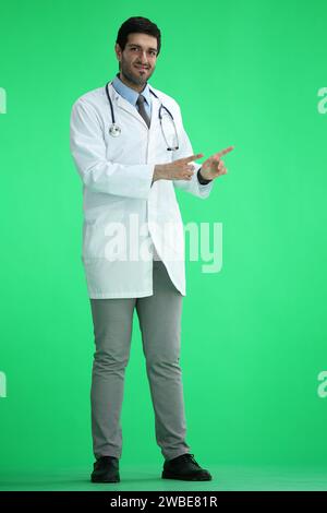 Männlicher Arzt auf grünem Hintergrund in vollem Wachstum zeigt zur Seite Stockfoto
