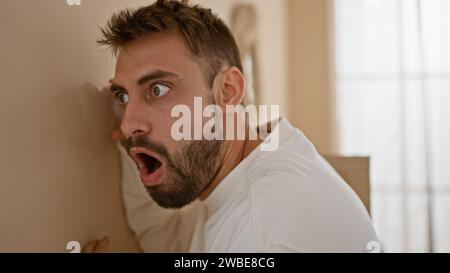 Neugieriger junger hispanischer Mann überrascht, als er zu Hause mit einem Glas durch die Wand lauschte Stockfoto