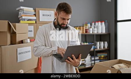 Der hübsche junge hispanische Mann, ein engagierter Freiwilliger, arbeitet unermüdlich und nutzt Laptop im Herzen eines belebten Wohltätigkeitszentrums. Sein entspanntes Gesicht und sein Bär Stockfoto