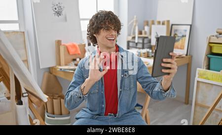 Ein fröhlicher junger Mann mit lockigem Haar, der in einem Kunststudio mit einem Tablet anruft, eine Jeansjacke und rote Kopfhörer trägt. Stockfoto