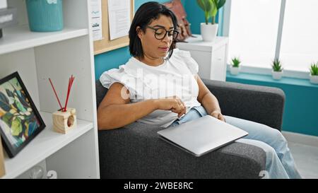 Hispanische Frau, die sich im modernen Büroinnenraum mit Laptop, lässig, Couch, Brille, besinnlich, Einrichtung, Business, Indoor, Erwachsener, Profi Stockfoto