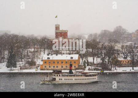 Ein Schneesturm in Stockholm, Schweden. Das Schloss Kastellet in Rot auf Kastellholmen mit schwedischer Flagge, vorbeifahrendes Pendlerboot. Stockfoto