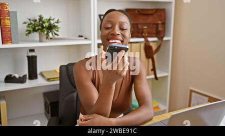 Fleißige afroamerikanische Geschäftsfrau, die mühelos schnelle Sprachnachrichten per Smartphone an ihrem eleganten Büroarbeitsplatz sendet Stockfoto