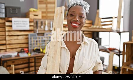 Lächelnde afroamerikanische Zimmerfrau genießt ihren Job mit Holzplanke in der Tischlerei Stockfoto