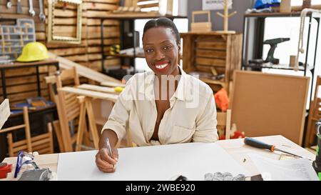 Schöne afroamerikanische Zimmerfrau lächelt und zeichnet in einer professionellen Holzwerkstatt des Handwerkers Stockfoto