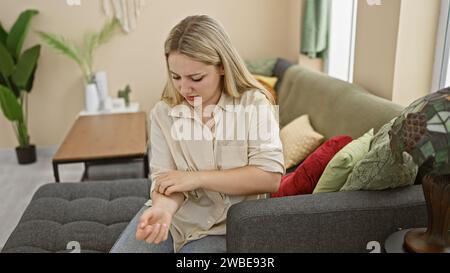 Eine junge, blonde Frau kratzt sich am Arm, während sie auf einer Couch in einem gemütlichen Wohnzimmer sitzt. Stockfoto