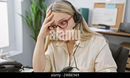 Eine gestresste junge blonde Frau mit Brille und Headset in einem Büro. Stockfoto