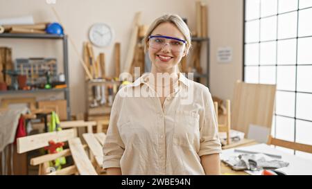 Eine lächelnde blonde Frau mit Schutzbrille steht selbstbewusst in einer gut beleuchteten Tischlerei. Stockfoto