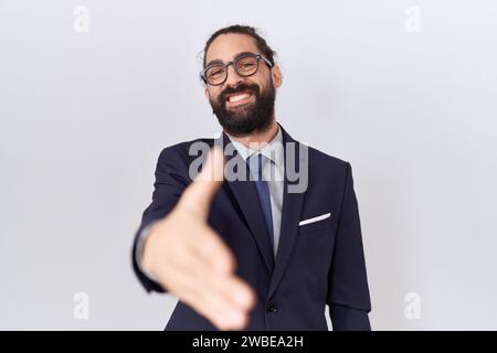 Hispanischer Mann mit Bart, der Anzug und Krawatte trägt und freundlich lächelt und Handshake als Begrüßung und Begrüßung anbietet. Erfolgreiche Geschäfte. Stockfoto