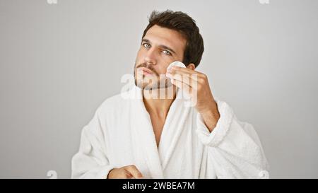 Schöner junger Mann im weißen Bademantel, der Gesichtspflege auf isoliertem Hintergrund anwendet Stockfoto