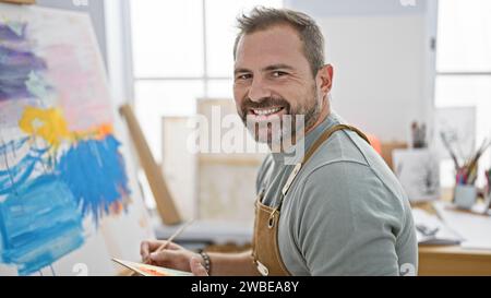 Hübscher reifer Mann mit grauen Haaren, mit Schürze, malt auf einer Leinwand in einem hellen Kunststudio. Stockfoto