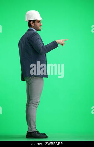 Ein Mann in einem Büroanzug, der einen Schutzhelm auf grünem Hintergrund trägt, zeigt zur Seite, im Profil Stockfoto