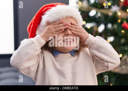 Entzückendes blondes Kleinkind, das die Augen bedeckt, mit Händen, die zu Hause am weihnachtsbaum stehen Stockfoto