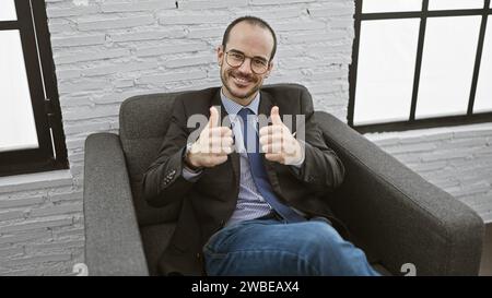 Selbstbewusster kahlköpfiger Mann mit Bart im Anzug, der die Daumen hochlegt, in moderner Büroumgebung Stockfoto