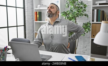 Ein reifer Mann mit grauem Bart erlebt Rückenschmerzen in einer modernen Büroumgebung und drückt Unbehagen aus. Stockfoto