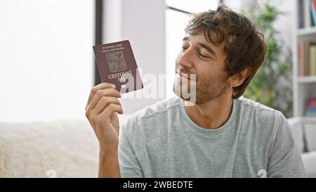 Selbstbewusster, bärtiger junger Mann, der auf seinem Wohnzimmer-Sofa sitzt, mit einem breiten Grinsen mit tschechischem Pass, voller Glück, bereit zum Einsteigen Stockfoto