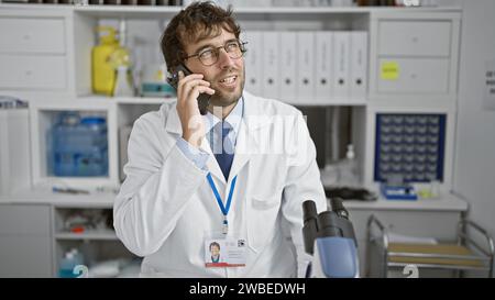 Ein hübscher junger blonder Wissenschaftler, der selbstbewusst lächelt und mit seinem Smartphone spricht und seine Laborarbeit mit losen Enden verbindet. Stockfoto