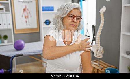 Reife Frau mit Schulterschmerzen im mit medizinischer Ausrüstung gefüllten Reha-Klinikraum. Stockfoto