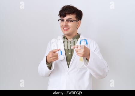 Junger, nicht binärer Mann mit Doktor Uniform und Stethoskop, der mit den Fingern auf die Kamera zeigt, mit glücklichem und lustigem Gesicht. Gute Energie und Vibes. Stockfoto
