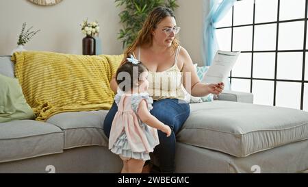 Zwanglose, entspannte Mutter und Tochter lesen liebevoll zusammen ein wichtiges Dokument auf ihrem bequemen Sofa und konzentrieren sich ruhig in ihrem gemütlichen Wohnraum Stockfoto