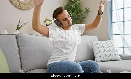 Ein fröhlicher junger hispanischer Mann entspannt sich mit Musik über Kopfhörer und tanzt auf einem Sofa in einer gemütlichen Wohnzimmerumgebung. Stockfoto