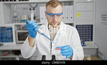 Ein fokussierter junger Mann im Laborkittel führt ein Experiment in einem Labor durch, indem er eine blaue Substanz in eine Petrischale pipettiert. Stockfoto