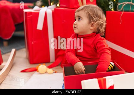 Entzückendes hispanisches Kleinkind, das zu Hause am weihnachtsbaum sitzt und ein Geschenk auspackt Stockfoto