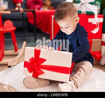 Entzückender hispanischer Junge, der zu Hause am weihnachtsbaum sitzt und ein Geschenk auspackt Stockfoto