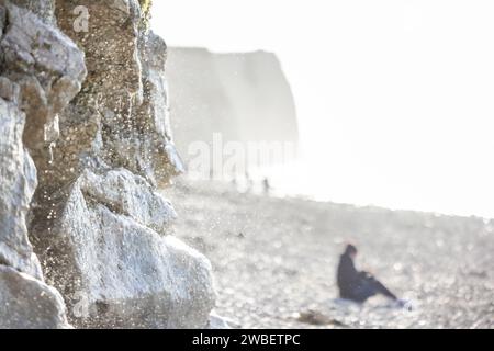 Dieses Bild zeigt einen natürlichen Wasserfall, der über eine sonnenverwöhnte felsige Klippe auf einen Kieselstrand stürzt. Der Fokus liegt auf den sprudelnden Wassertröpfchen, die das Sonnenlicht auffangen und einen glitzernden Effekt vor dem unscharfen Hintergrund erzeugen. In der Ferne sitzt eine Einzelfigur am Strand, die der Szene ein Gefühl von Größe und Einsamkeit verleiht. Die Klippe im Hintergrund ist in hellem Sonnenlicht getaucht, was die Textur der Felsen und die nebelige Qualität des Wassersprühens verbessert. Schimmernde Cliffside-Kaskade. Hochwertige Fotos Stockfoto