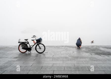 Frau, die einen Vogel an einem kalten Tag mit ihrem Fahrrad auf einem Stand in der Nähe fotografiert Stockfoto