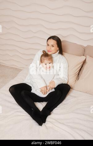 Familienfoto einer hübschen Mutter mit langen dunklen Haaren, die einen gestrickten weißen Pullover trägt und mit ihrer kleinen süßen Tochter in einem gemütlichen Bett posiert Stockfoto