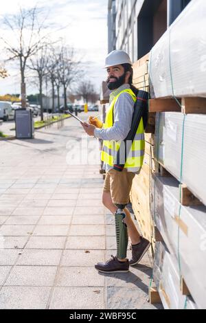 Vertikales Seitenporträt eines Ingenieurs mit Beinprothetik, der auf einer Baustelle im Freien arbeitet Stockfoto