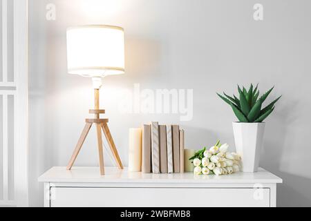 Glühende Lampe mit Büchern, Tulpen und Zimmerpflanzen auf der Kommode im Zimmer Stockfoto