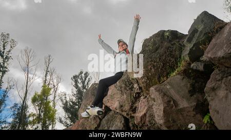 Schöne junge lateinamerikanische Frau, die auf Steinen auf einem Berg sitzt, mit erhobenen Armen an einem bewölkten Tag Stockfoto