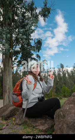 Schöne junge lateinamerikanische Frau, die auf Steinen auf dem Gipfel eines Berges sitzt, auf die Kamera blickt und mit ihren beiden Händen ein Siegeszeichen macht Stockfoto