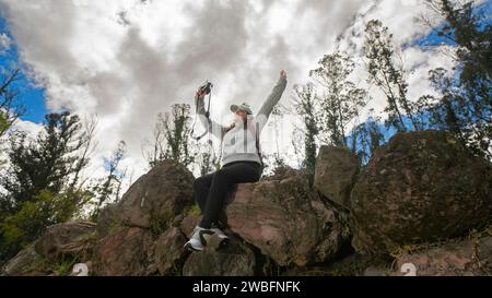 Schöne junge lateinamerikanische Frau, die auf Steinen auf einem Berg sitzt und an einem bewölkten Tag ein Selfie mit ihrer kleinen Kamera macht Stockfoto